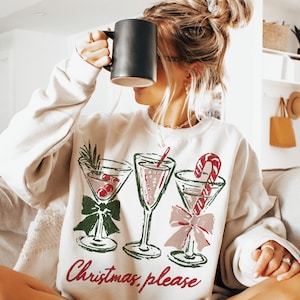 May include: Cream-colored sweatshirt with a festive cocktail design. The graphic features three martini glasses with red and green accents, and a candy cane. The text "Christmas, please" is printed in red.