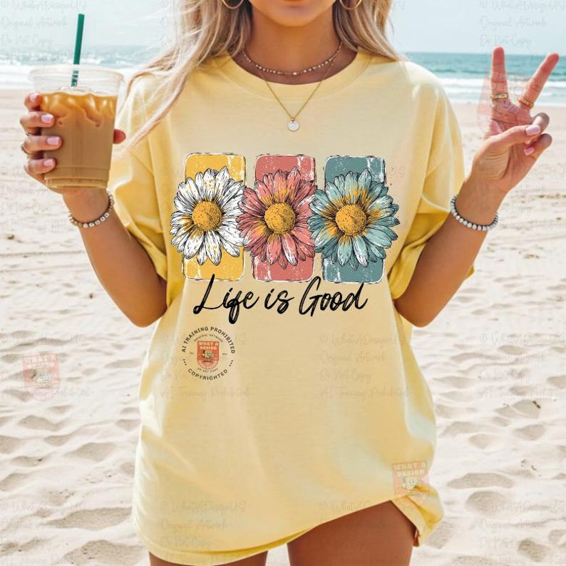 May include: A light yellow t-shirt featuring three daisy flowers in yellow, pink, and blue rectangular frames. The text "Life is Good" is printed below the flowers. The shirt is worn on a beach.