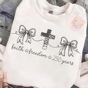 May include: White sweatshirt with a black line drawing of two bows and a cross with an American flag design. The words "faith freedom 250 years" are written in a script font below the drawings.