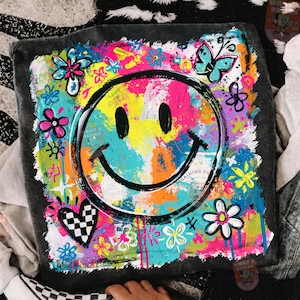 May include: A square, gray cushion with a vibrant, colorful design. The design features a large, black smiley face surrounded by flowers, butterflies, and a checkered heart. The background is a mix of bright colors including pink, yellow, and blue.