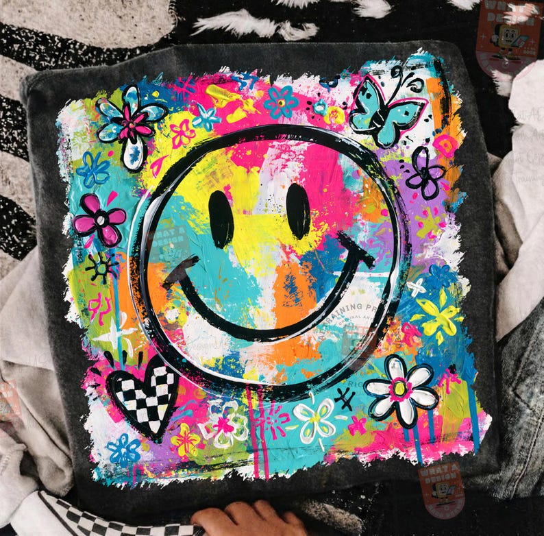 May include: A square, gray cushion with a vibrant, colorful design. The design features a large, black smiley face surrounded by flowers, butterflies, and a checkered heart. The background is a mix of bright colors including pink, yellow, and blue.