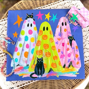 May include: A blue t-shirt featuring three ghosts wearing patterned sheets. The left ghost has colorful polka dots, the center ghost has orange pumpkins, and the right ghost has pink flowers. Each ghost is accompanied by a black cat, with colorful stars.
