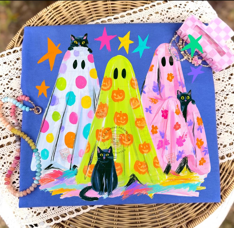 May include: A blue t-shirt featuring three ghosts wearing patterned sheets. The left ghost has colorful polka dots, the center ghost has orange pumpkins, and the right ghost has pink flowers. Each ghost is accompanied by a black cat, with colorful stars.