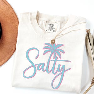 May include: A white t-shirt with a blue and pink palm tree graphic and the word "Salty" in a blue and pink script font.