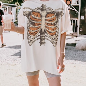 May include: Cream-colored oversized t-shirt featuring a skeletal rib cage design with orange jack-o'-lanterns inside. The graphic tee is perfect for Halloween or spooky season. The shirt is a relaxed fit.