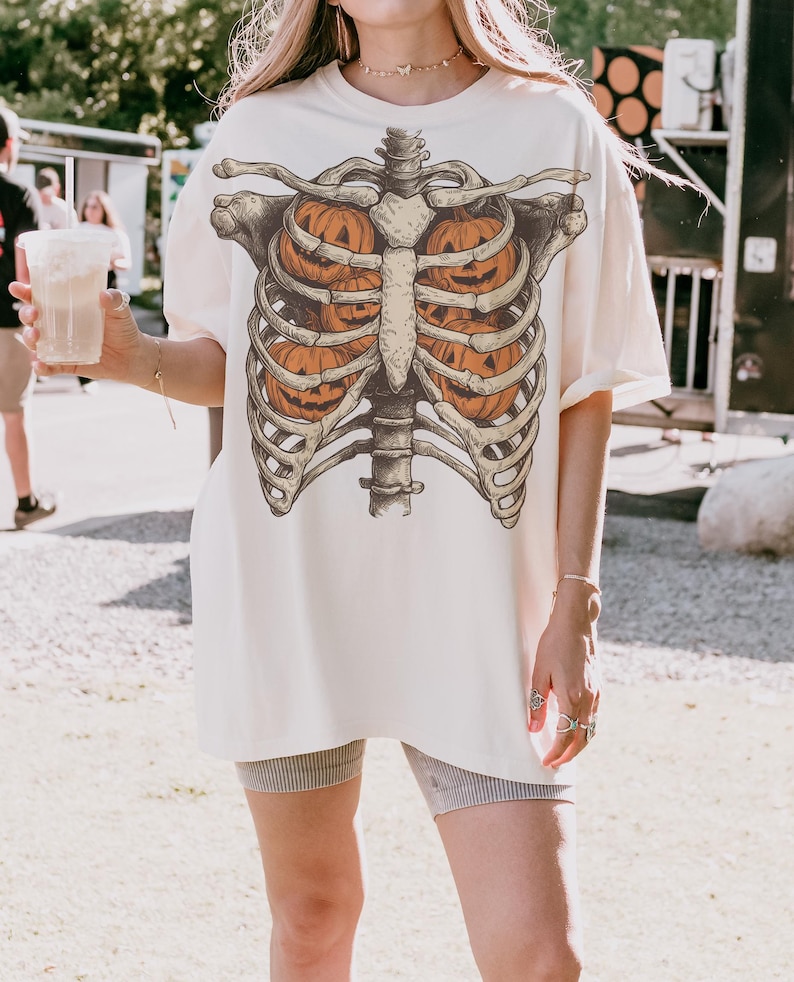 May include: Cream-colored oversized t-shirt featuring a skeletal rib cage design with orange jack-o'-lanterns inside. The graphic tee is perfect for Halloween or spooky season. The shirt is a relaxed fit.