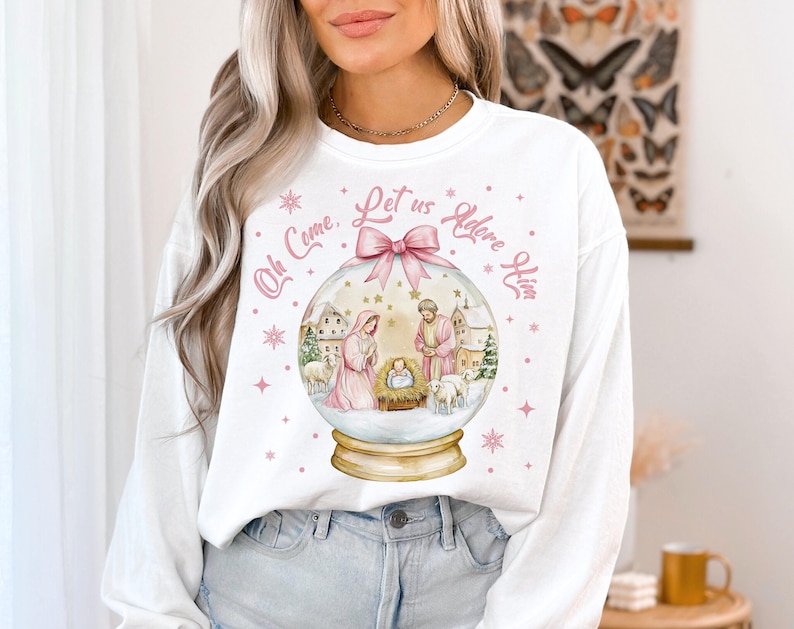 K&ouml;nnte beinhalten: Wei&szlig;er Langarm-Pullover mit einem rosa-wei&szlig;en Schneekugel-Design. Die Schneekugel zeigt eine Weihnachtskrippe mit Maria, Josef und dem Jesuskind. Der Text "Oh Come, Let us Adore Him" ist um die Schneekugel herum geschrieben.
