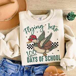 May include: A cream-coloured sweatshirt with the text "Flying Into 100 Days of School" and a cartoon illustration of a duck wearing sunglasses and a hat, flying with a backpack.