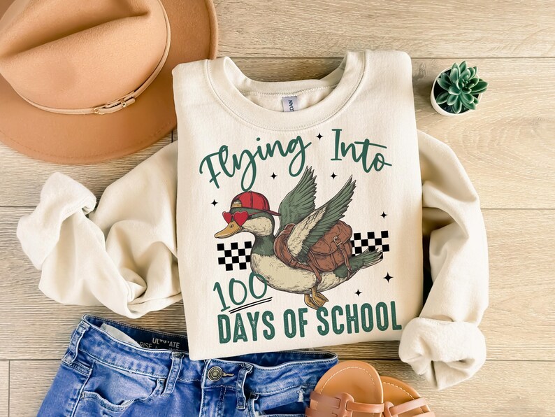 May include: A cream-coloured sweatshirt with the text "Flying Into 100 Days of School" and a cartoon illustration of a duck wearing sunglasses and a hat, flying with a backpack.