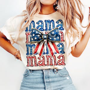 May include: Cream-colored t-shirt with the word "Mama" in blue and red, with a patriotic bow graphic. The bow features a blue star pattern and red and white stripes, evoking an American theme.