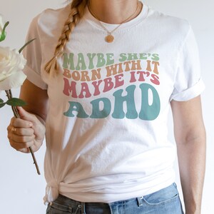 May include: White t-shirt with the text "MAYBE SHE'S BORN WITH IT MAYBE IT'S ADHD" in retro-style lettering. The text is in shades of green, orange, pink, and teal. The shirt has rolled-up sleeves.