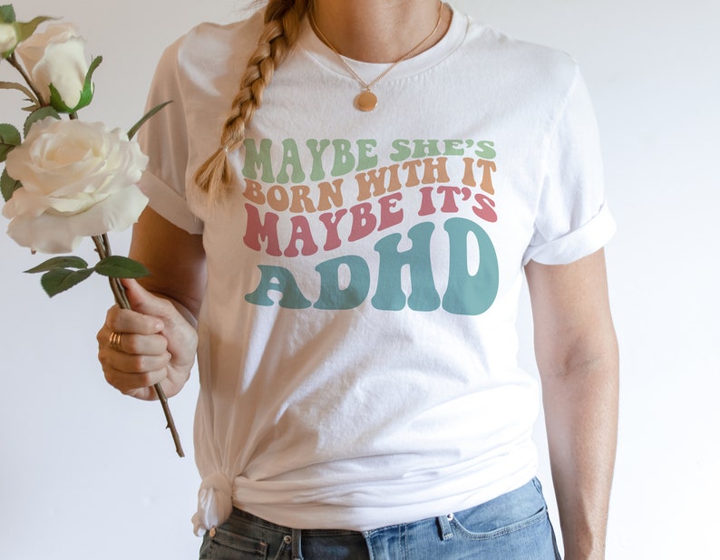 May include: White t-shirt with the text "MAYBE SHE'S BORN WITH IT MAYBE IT'S ADHD" in retro-style lettering. The text is in shades of green, orange, pink, and teal. The shirt has rolled-up sleeves.