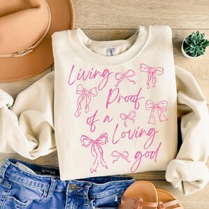 May include: A cream-coloured sweatshirt with the text "Living Proof of a Loving Good" in pink script. The text is surrounded by pink bow illustrations.