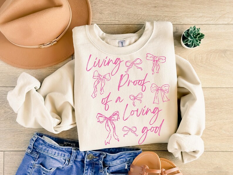 May include: A cream-coloured sweatshirt with the text "Living Proof of a Loving Good" in pink script. The text is surrounded by pink bow illustrations.