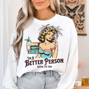 May include: White long-sleeve sweatshirt with a graphic print of a woman in a polka dot swimsuit and palm trees. The text "I'm a Better Person When I'm Tan" is printed below the graphic.
