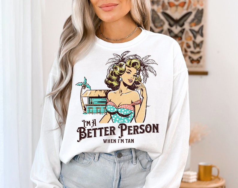 May include: White long-sleeve sweatshirt with a graphic print of a woman in a polka dot swimsuit and palm trees. The text "I'm a Better Person When I'm Tan" is printed below the graphic.