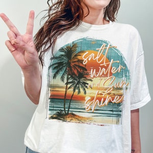 May include: White t-shirt with a graphic print of a sunset over the ocean with palm trees and the text "salt water and sun shine".