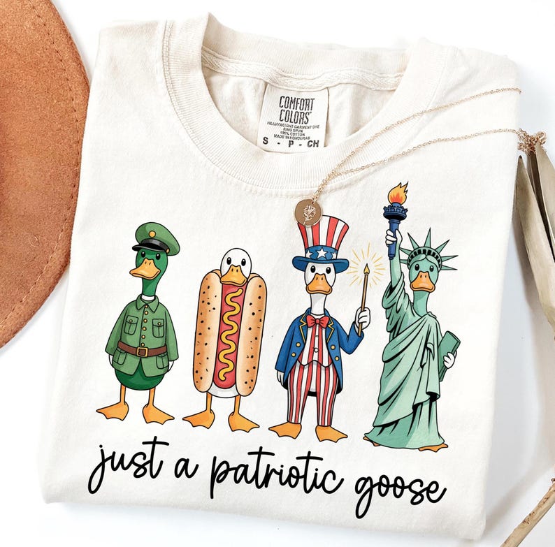 May include: Cream-colored t-shirt featuring a whimsical design of patriotic geese. The geese are dressed as a soldier, a hot dog, Uncle Sam, and the Statue of Liberty. The text "just a patriotic goose" is written in a script font.