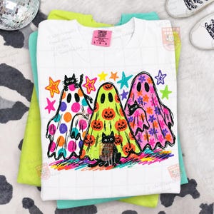 K&ouml;nnte beinhalten: Ein wei&szlig;es T-Shirt mit einer Grafik von drei Geistern in verschiedenen Mustern: einer mit orangefarbenen und pinken Punkten, einer mit orangefarbenen K&uuml;rbissen und einer mit pinken Blumen. Drei schwarze Katzen sind zwischen den Geistern und bunten Sternen.