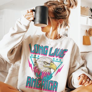 May include: A white sweatshirt with a graphic print of an eagle with the text "Long Live America" in pink, blue, and teal.