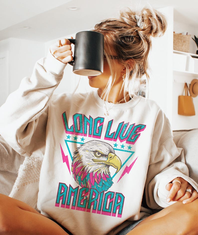 May include: A white sweatshirt with a graphic print of an eagle with the text "Long Live America" in pink, blue, and teal.