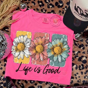 May include: A bright pink t-shirt featuring three large daisy flower graphics in white, pink, and blue, each with a yellow center. The shirt has the phrase "Life is Good" printed in black script below the flowers.