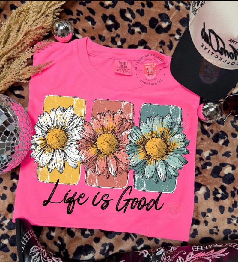 May include: A bright pink t-shirt featuring three large daisy flower graphics in white, pink, and blue, each with a yellow center. The shirt has the phrase "Life is Good" printed in black script below the flowers.