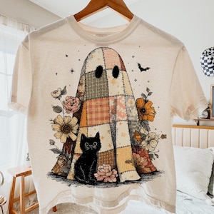 Könnte beinhalten: Cremefarbenes T-Shirt mit einem Patchwork-Geist-Design mit einer schwarzen Katze, Blumen und einer Fledermaus. Der Geist besteht aus verschiedenen gemusterten Stoffen in Beige-, Orange- und Grüntönen. Das Shirt hängt an einem Holzbügel.