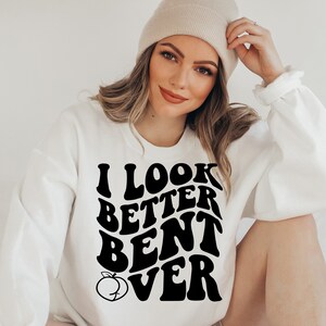 May include: White sweatshirt with black text that reads "I look better bent over" with a peach graphic. The model is wearing a beige beanie.