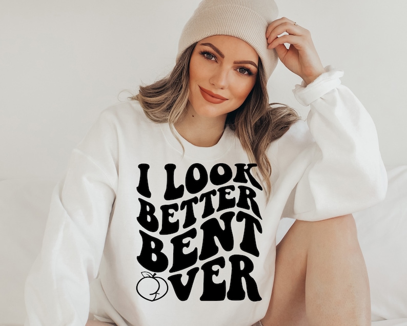 May include: White sweatshirt with black text that reads "I look better bent over" with a peach graphic. The model is wearing a beige beanie.