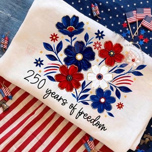 May include: A white decorative cloth with a floral design in red, white, and blue. The flowers are arranged in a bouquet with gold accents. The text "250 years of freedom" is written in black script.