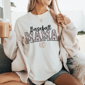 May include: White sweatshirt with the text "Baseball Mama" in black lettering with a red baseball stitch design. A white baseball with red stitching is below the text.