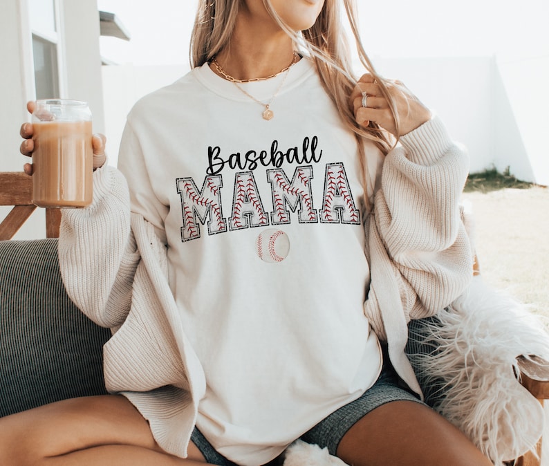 May include: White sweatshirt with the text "Baseball Mama" in black lettering with a red baseball stitch design. A white baseball with red stitching is below the text.