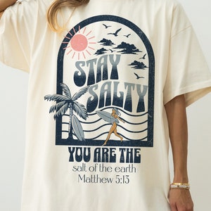 May include: A cream colored t-shirt with a graphic print of a sun, clouds, palm trees, a surfer, and the text "Stay Salty" and "You are the salt of the earth Matthew 5:13".