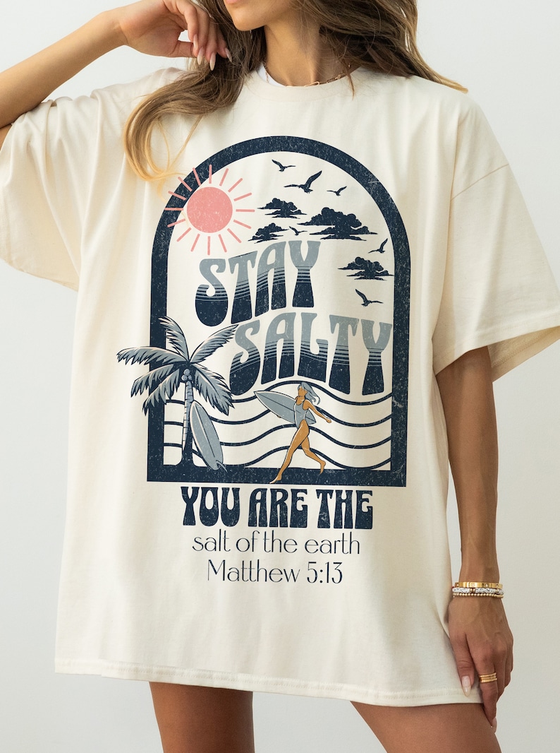 May include: A cream colored t-shirt with a graphic print of a sun, clouds, palm trees, a surfer, and the text "Stay Salty" and "You are the salt of the earth Matthew 5:13".