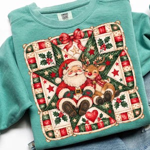 May include: A seafoam green crewneck sweatshirt featuring a graphic of Santa Claus and a reindeer sitting together on a large green star. The design includes red bows, holly berries, and a red heart, all framed by a patchwork-style border.