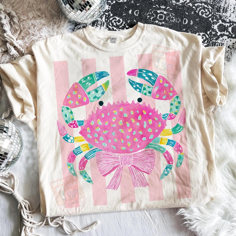 May include: Cream-colored t-shirt featuring a colorful crab design. The crab is pink with blue, green, and yellow accents, and a pink bow. The background has pink vertical stripes. The shirt is from Comfort Colors.