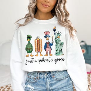 May include: White sweatshirt with a graphic of four cartoon geese dressed as patriotic symbols: a soldier, a hot dog, Uncle Sam, and the Statue of Liberty. The text "just a patriotic goose" is written below the geese.
