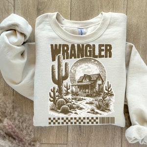 Pode incluir: Uma camisa de moletom bege com uma estampa gr&aacute;fica marrom de uma casa de estilo western, um cacto e a palavra "WRANGLER".