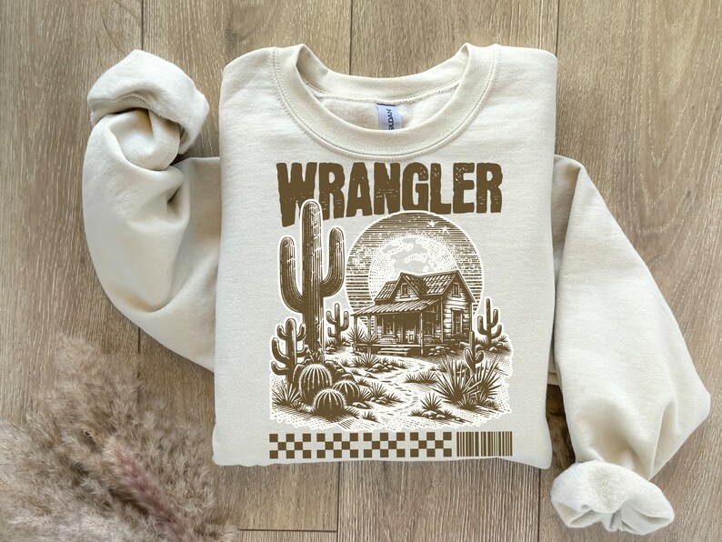 Pode incluir: Uma camisa de moletom bege com uma estampa gr&aacute;fica marrom de uma casa de estilo western, um cacto e a palavra "WRANGLER".