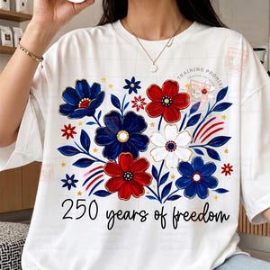 May include: White t-shirt featuring a floral design in red, white, and blue, with gold accents. The design includes the text "250 years of freedom" in a script font. The shirt has a crew neck and short sleeves.