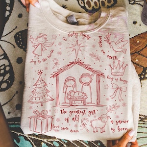 K&ouml;nnte beinhalten: Ein beiges Sweatshirt mit einem roten Weihnachtsmotiv. Das Design umfasst einen Engel, einen Weihnachtsbaum, eine Krippe, ein Geschenk, ein Schaf und die S&auml;tze "Joy to the World", "Holy Night", "Peace on Earth", "King of Kings", "The Greatest Gift of All", "Love Came Down" und "A Savior is Born".