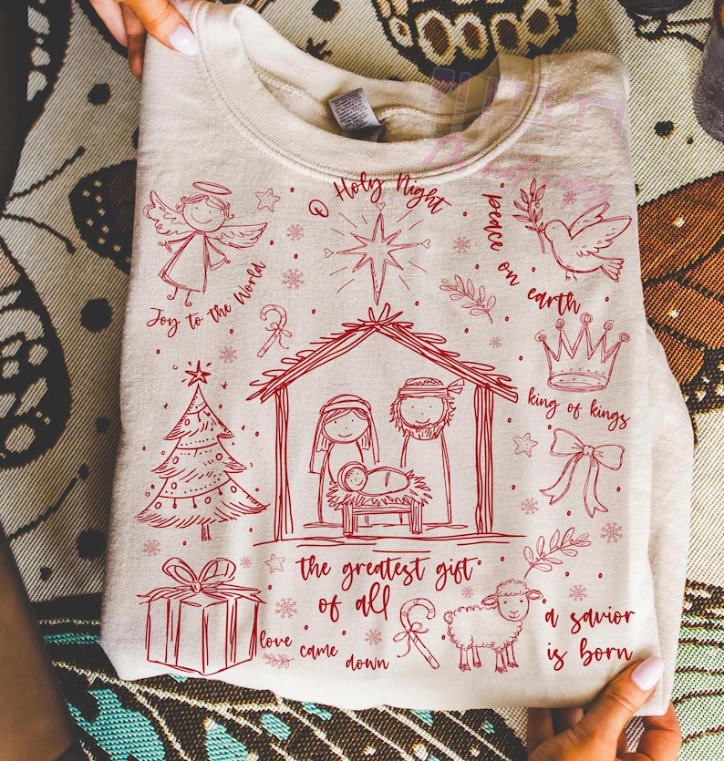K&ouml;nnte beinhalten: Ein beiges Sweatshirt mit einem roten Weihnachtsmotiv. Das Design umfasst einen Engel, einen Weihnachtsbaum, eine Krippe, ein Geschenk, ein Schaf und die S&auml;tze "Joy to the World", "Holy Night", "Peace on Earth", "King of Kings", "The Greatest Gift of All", "Love Came Down" und "A Savior is Born".