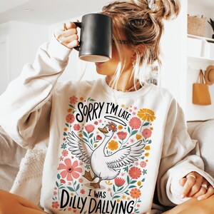 May include: White sweatshirt with a floral design and a goose illustration. The text on the sweatshirt reads "Sorry I'm Late I Was Dilly Dallying".
