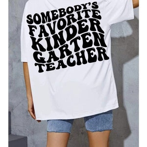 May include: White t-shirt with the text "SOMEBODY'S FAVORITE KINDERGARTEN TEACHER" in large, black, bubble letters. The shirt is a casual style, with short sleeves and a relaxed fit. The person is wearing denim shorts.