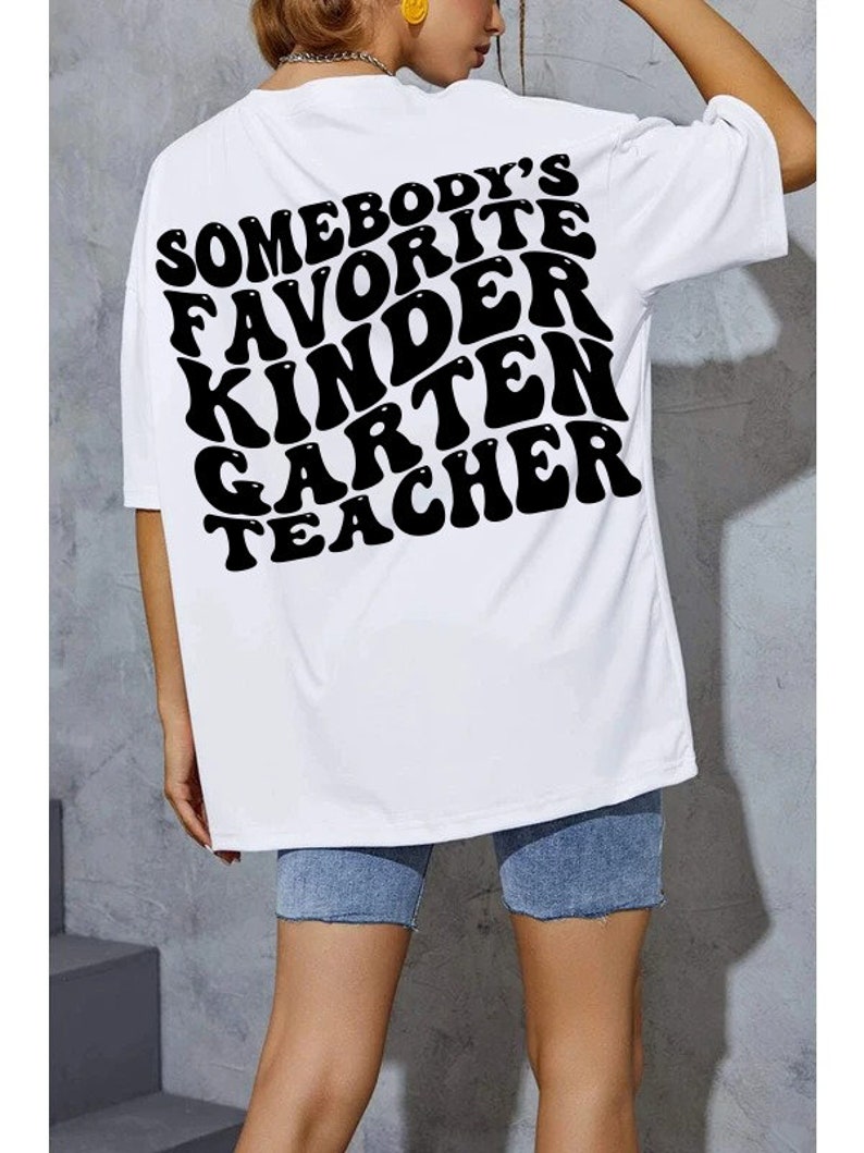 May include: White t-shirt with the text "SOMEBODY'S FAVORITE KINDERGARTEN TEACHER" in large, black, bubble letters. The shirt is a casual style, with short sleeves and a relaxed fit. The person is wearing denim shorts.