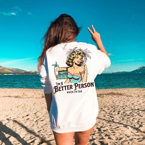 May include: White sweatshirt with a graphic print of a woman in a blue and white polka dot swimsuit with a palm tree behind her. The text on the shirt reads "I'm a Better Person When I'm Tan."