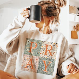 May include: A cream-colored crewneck sweatshirt featuring a square graphic divided into four quadrants. Each quadrant displays a floral pattern in muted tones of green, pink, and white, with the large orange letters P, R, A, and Y overlaid on top.