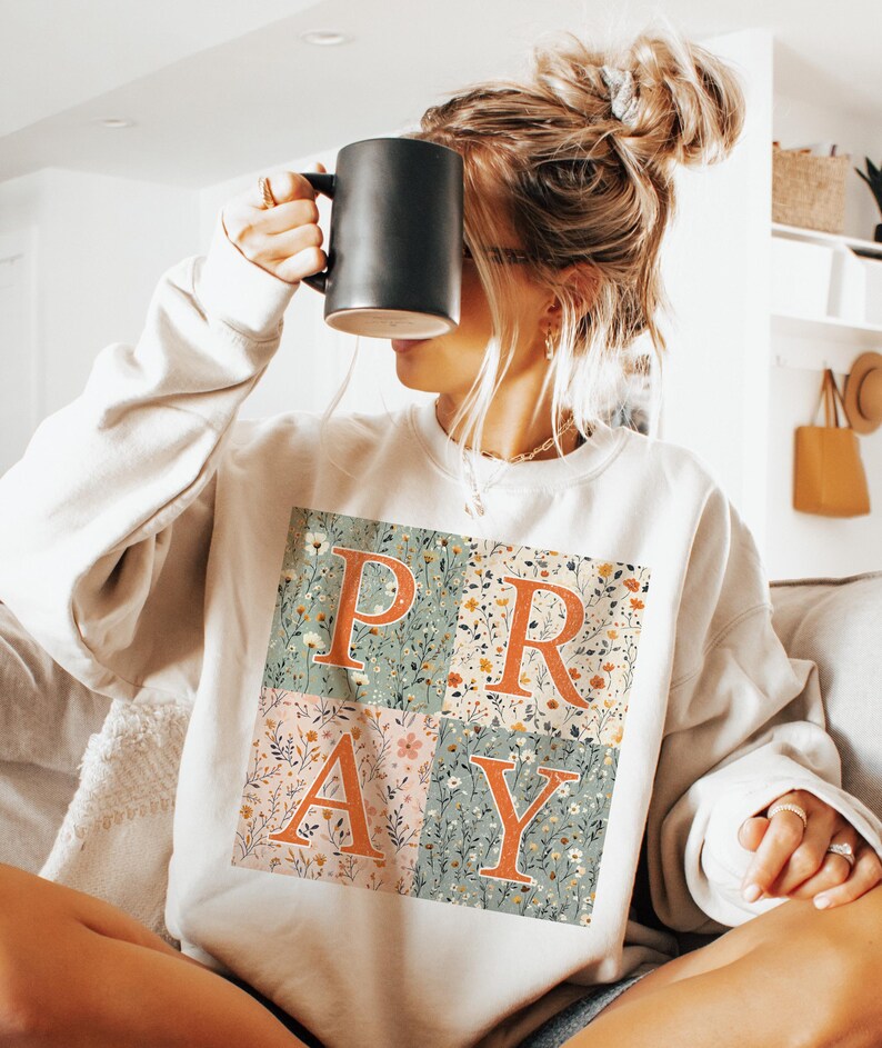 May include: A cream-colored crewneck sweatshirt featuring a square graphic divided into four quadrants. Each quadrant displays a floral pattern in muted tones of green, pink, and white, with the large orange letters P, R, A, and Y overlaid on top.