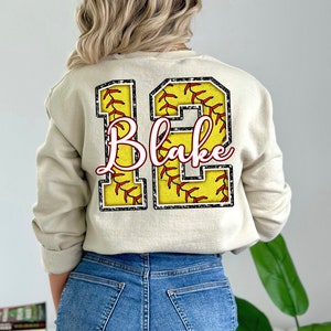 May include: A beige sweatshirt with a large number "12" printed on the back. The number is made up of two softball designs with red stitching and a black outline. The name "Blake" is written in pink cursive inside the number.